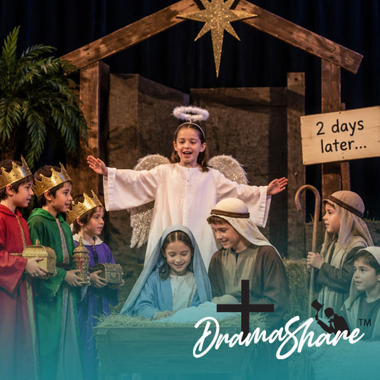 A children's nativity scene performance with a young girl dressed as an angel standing above a baby in a manger, surrounded by children in royal and shepherd costumes. A wooden sign reads '2 days later.